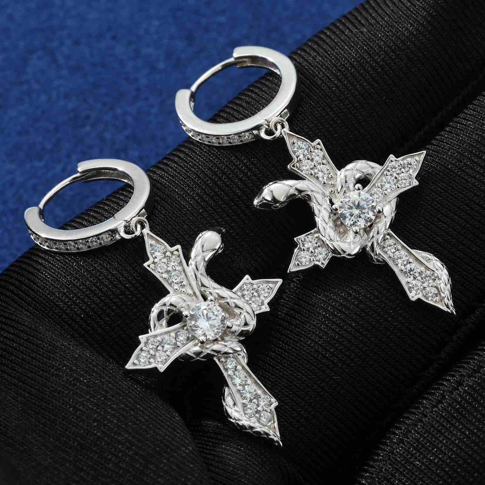 White gold snake cross earrings displayed on black gloves, emphasizing moissanite sparkle and metallic sheen through high contrast.